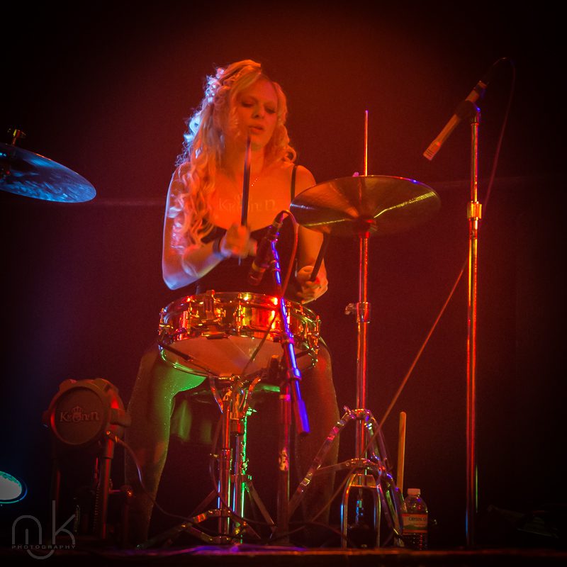 Drummer performing on stage with red lighting.